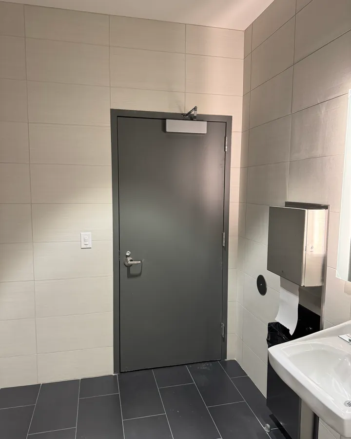 New gray commercial steel door with an overhead closer and lever handle in a tiled restroom